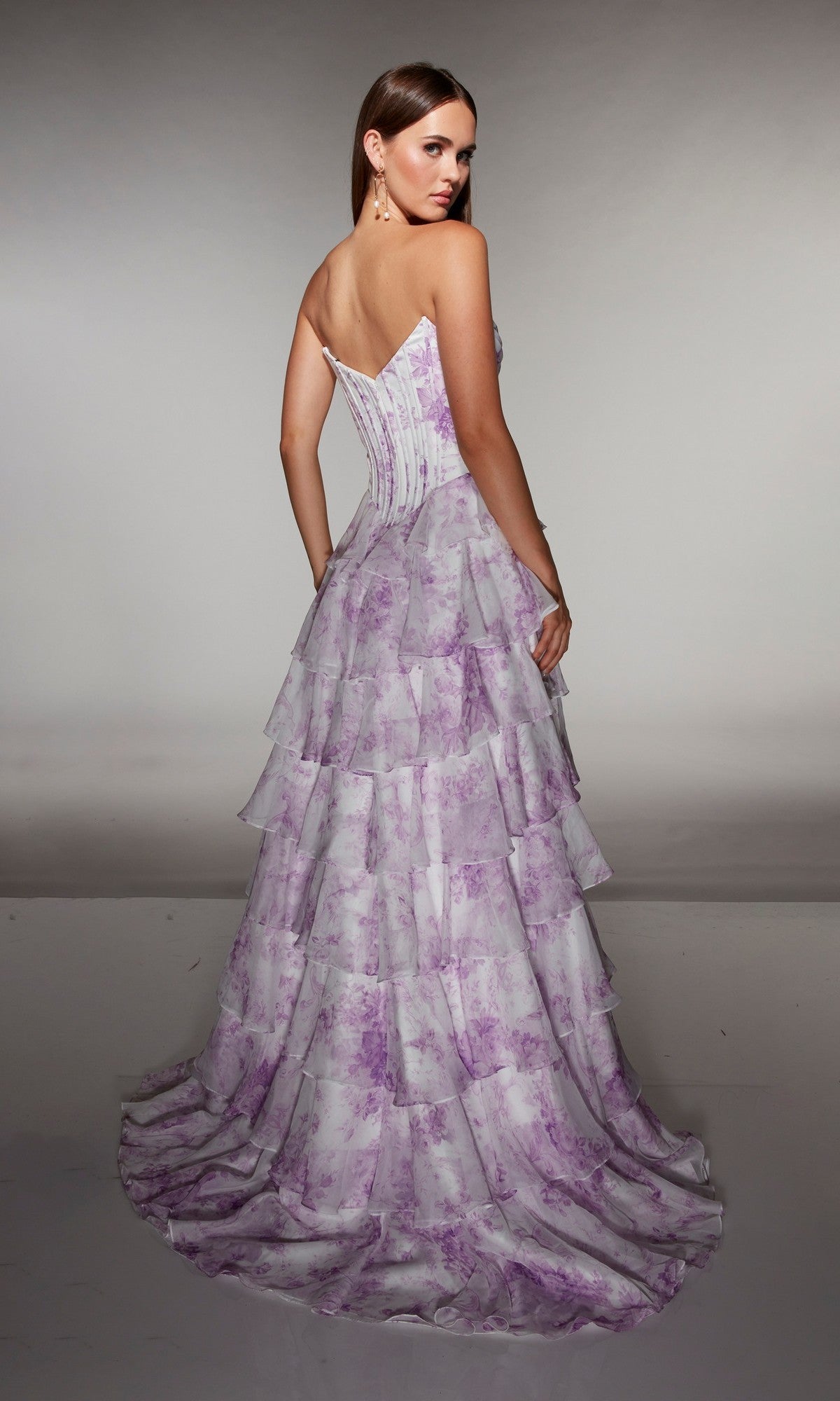 A woman faces away, wearing the Alyce 62192 long prom dress featuring a strapless corset bodice and tiered floor-length chiffon ruffle skirt with a lavender floral pattern, set against a neutral background.